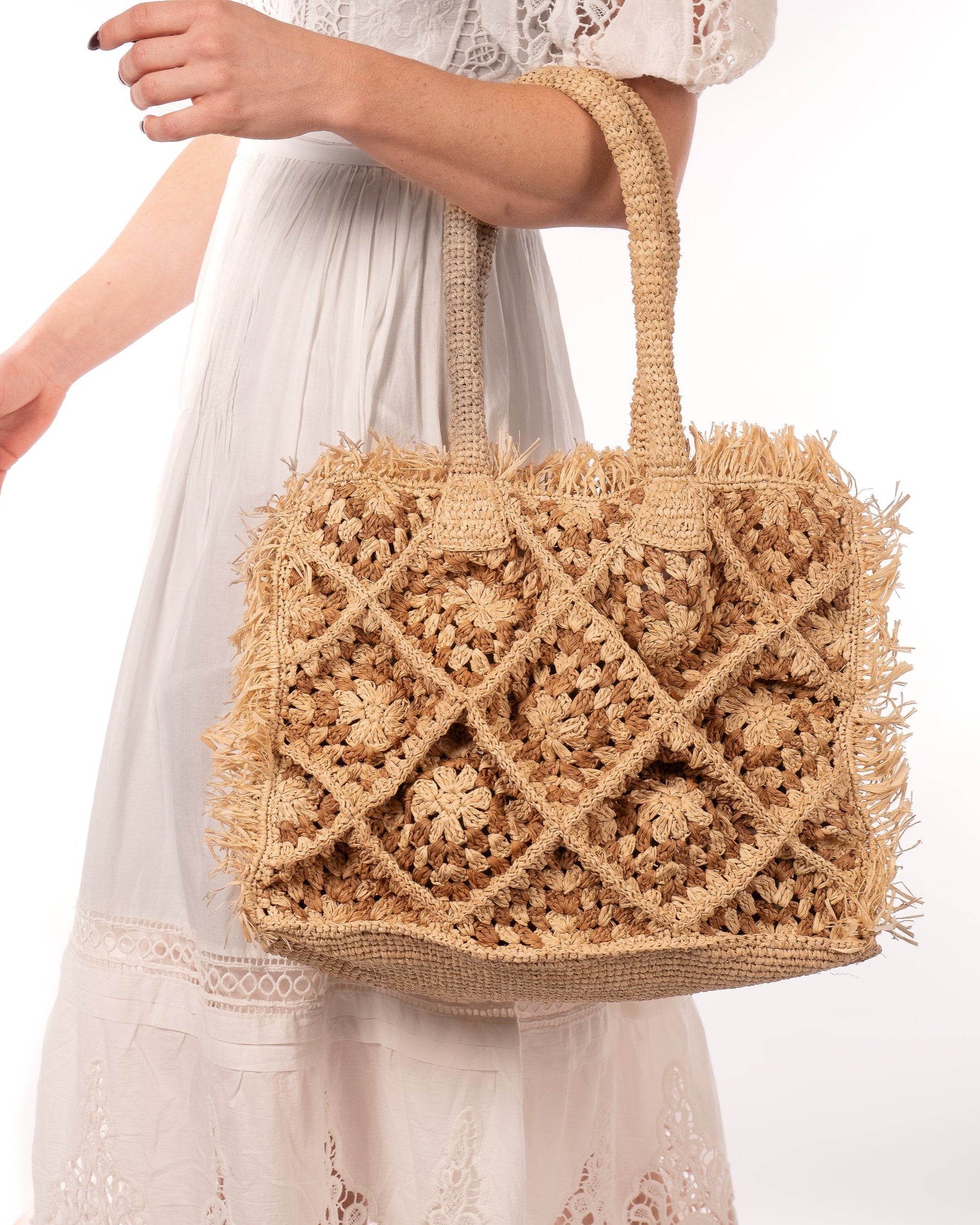 Model wearing Leo Crochet Granny Square Pattern Handbag with Fringe Edge handmade woven raffia palm fiber in a two tone cappuccino light/brown/beige and natural straw color multicolor pattern of woven granny squares oriented on the diagonal with natural raw fringe edging and handles basket bag - Shebobo