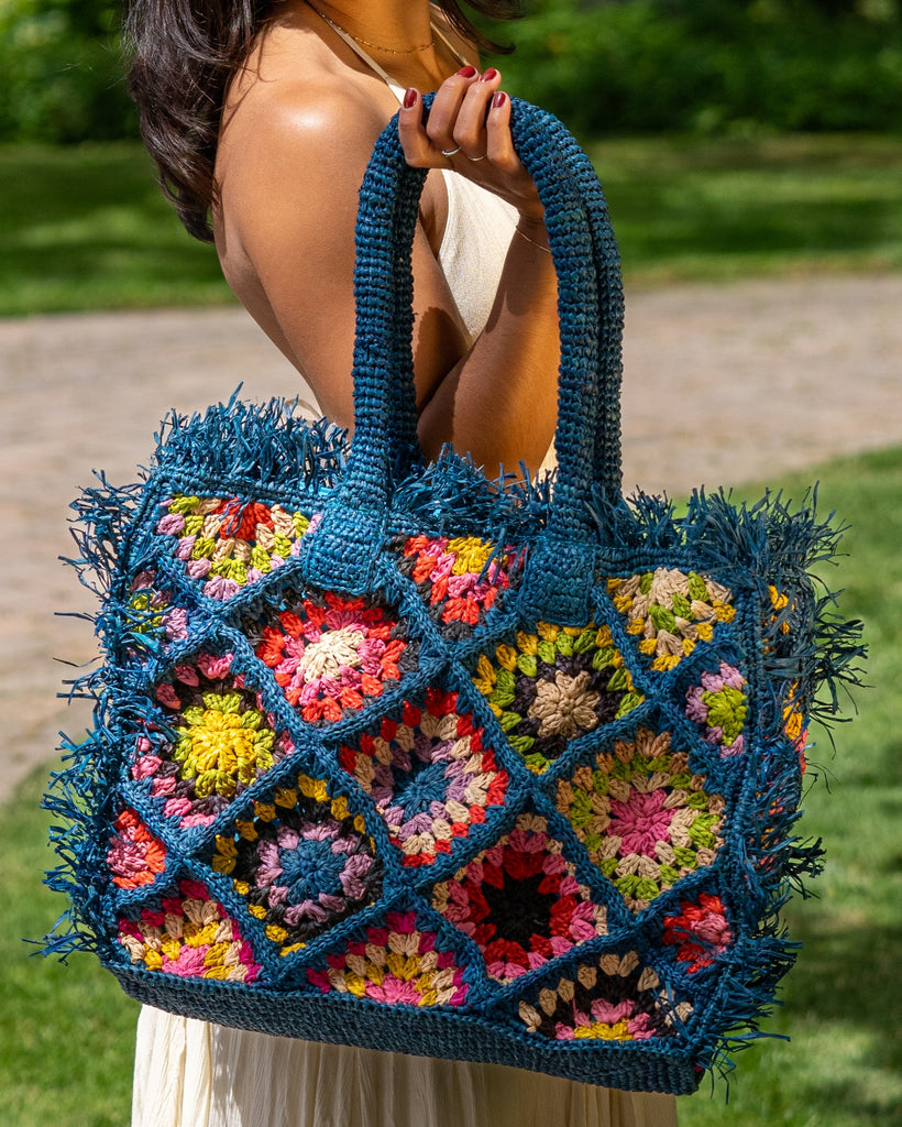 Model wearing Leo Crochet Granny Square Pattern Handbag with Fringe Edge handmade woven raffia palm fiber in a turquoise blue, coral red/orange, yellow, lime green, pink, black, natural straw color, etc. multicolor pattern of woven granny squares oriented on the diagonal with natural raw fringe edging and handles basket bag - Shebobo