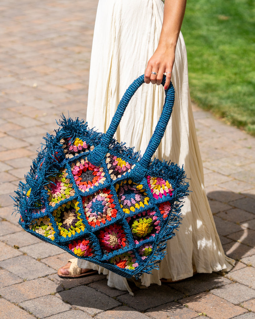 Model wearing Leo Crochet Granny Square Pattern Handbag with Fringe Edge handmade woven raffia palm fiber in a turquoise blue, coral red/orange, yellow, lime green, pink, black, natural straw color, etc. multicolor pattern of woven granny squares oriented on the diagonal with natural raw fringe edging and handles basket bag - Shebobo