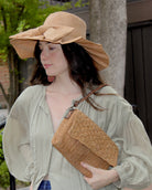 Model wearing Lilly 5" Brim Fan Straw Sun Hat with Big Bow Cappuccino handmade loomed natural raffia palm fiber in a solid hue of cappuccino tea/light/brown with pleated wide brim and oversized bow embellishment - Shebobo (with Anna Petal Pattern Crochet Straw Over Shoulder Envelope Clutch Purse)