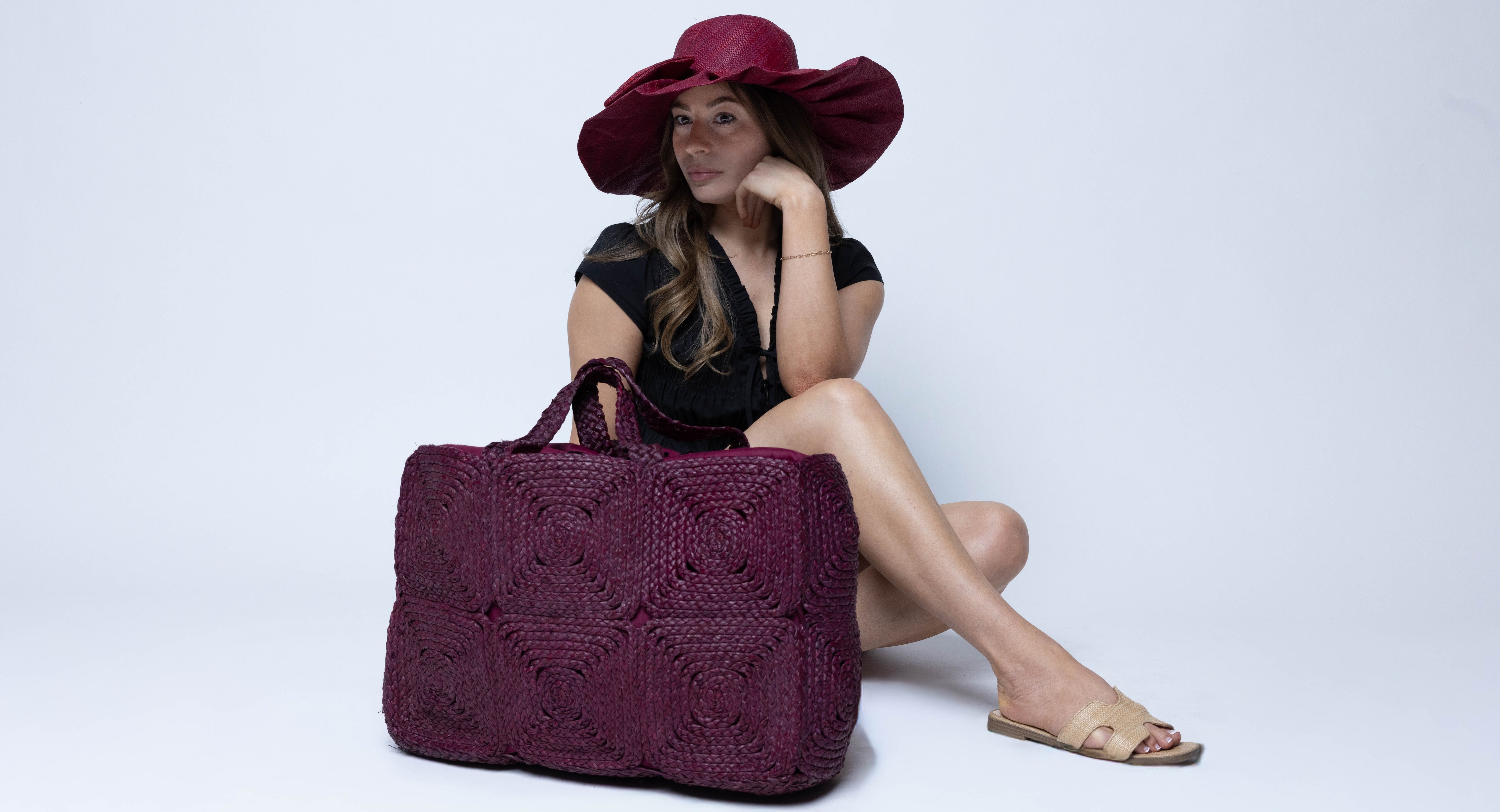 Model wearing Gemma Crochet Disc Rectangle Large Straw Basket Handbag handmade braided natural raffia palm fiber in a solid hue of Umberwine burgundy/bordeaux/dark/purple/red with subtle square disc pattern around the outside of the bag in two horizontal bands of squares creating a geometric pattern and matching woven handles beach bag purse - Shebobo (with Lilly 5" Brim Fan Pleated Straw Sun Hats with Big Bow)