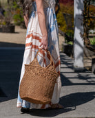 Model wearing Luca Macrame Diamonds Crochet Straw Handbag handmade knotted natural raffia palm fiber in a solid hue of Cappuccino tea/light/brown in a multi-texture circular pattern of concentric spiral links on the base, cross-hatch x pattern up 2/3rds of the bag, and geometric diamond flower pattern around the top third, with diamond pattern on the base of the shoulder strap handles with tubular loop handle tops - Shebobo