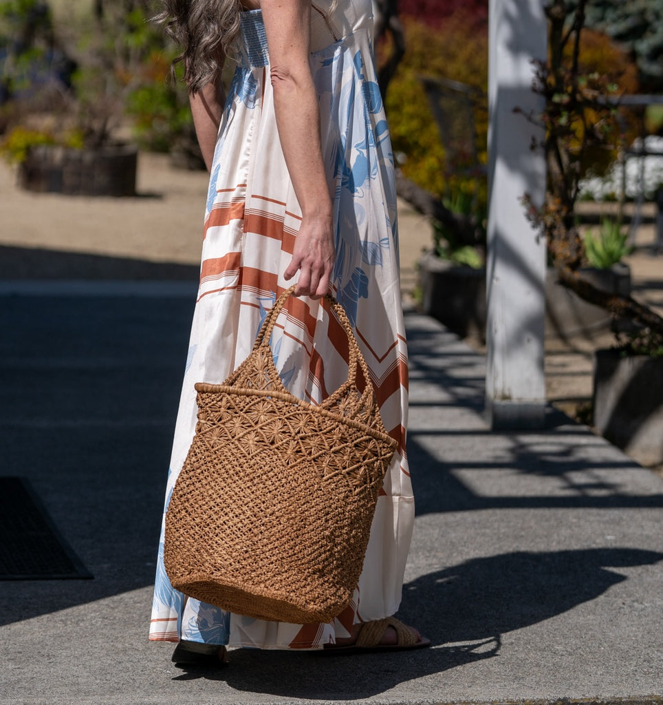 Model wearing Luca Macrame Diamonds Crochet Straw Handbag handmade knotted natural raffia palm fiber in a solid hue of Cappuccino tea/light/brown in a multi-texture circular pattern of concentric spiral links on the base, cross-hatch x pattern up 2/3rds of the bag, and geometric diamond flower pattern around the top third, with diamond pattern on the base of the shoulder strap handles with tubular loop handle tops - Shebobo