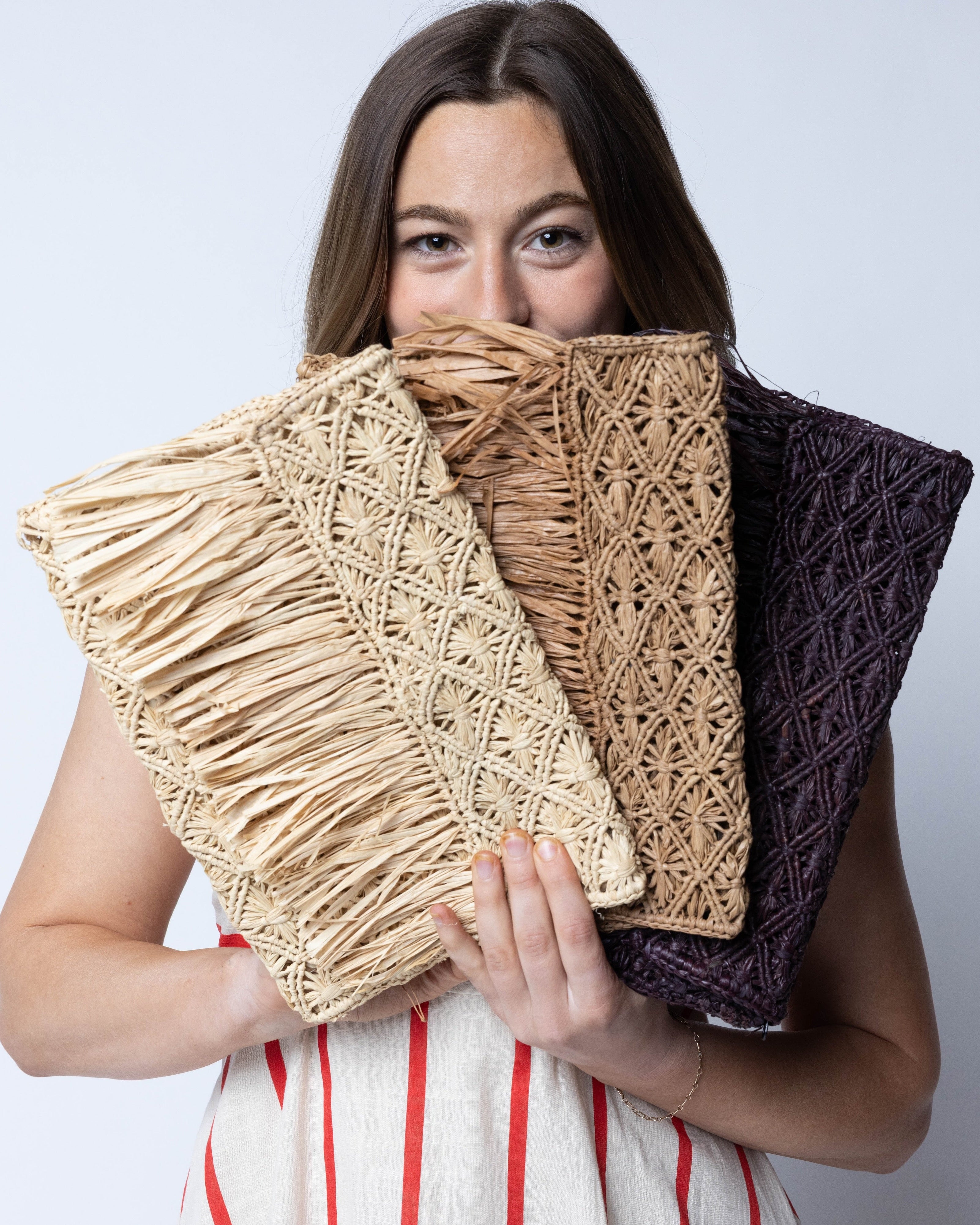 Model wearing Multiple color options Luli Macrame Diamond and Crochet Straw Envelope Clutch Purse with Raw Fringe Trim Edge handmade macrame and crochet natural raffia palm fiber in a solid hue of Mocha, Natural, or Cappuccino in a diagonal geometric pattern of starburst or flower inside diamonds with hand brushed raw fringe trim edge along the top flap of the envelope design handbag  - Shebobo