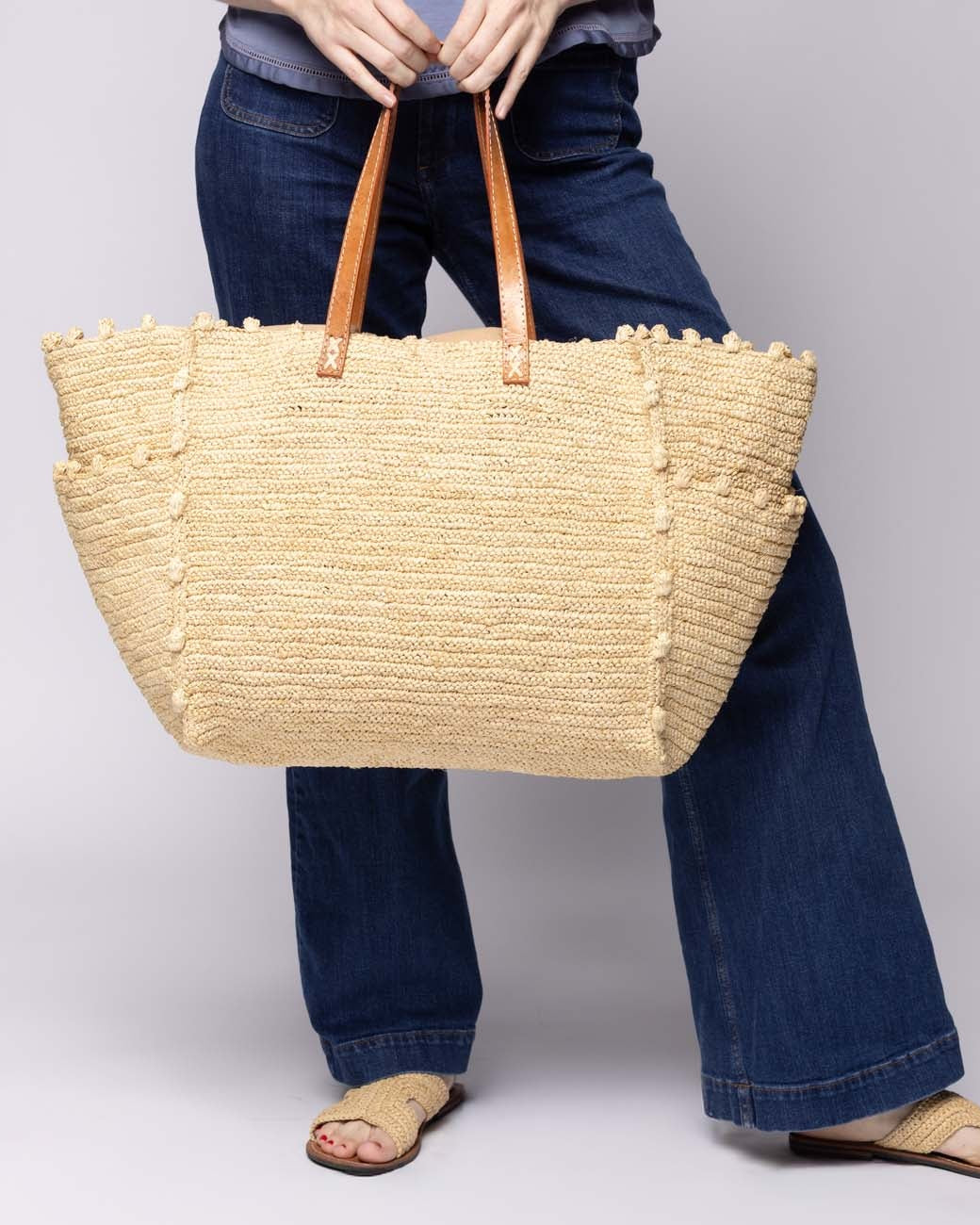 Model wearing Madeline Natural XL Crochet Raffia Straw Basket Shoulder Bag with Pockets handmade raffia palm fiber in a solid hue of natural straw color with crochet horizontal subtle band texture, round boucle nub edges, oversized external pockets, and leather handles - Shebobo