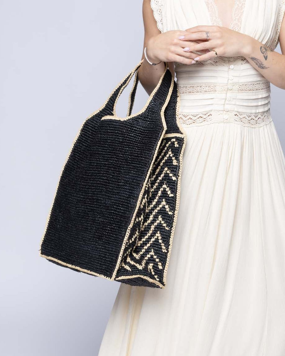 Model wearing Side view Mamie Zigzag Pinstripe XL Crochet Straw Basket Handbag handmade crochet natural raffia palm fiber in a solid hue of black purse beach bag with contrasting natural straw color zigzag pinstripe pattern on the sides of the bag and matching outer edge - Shebobo