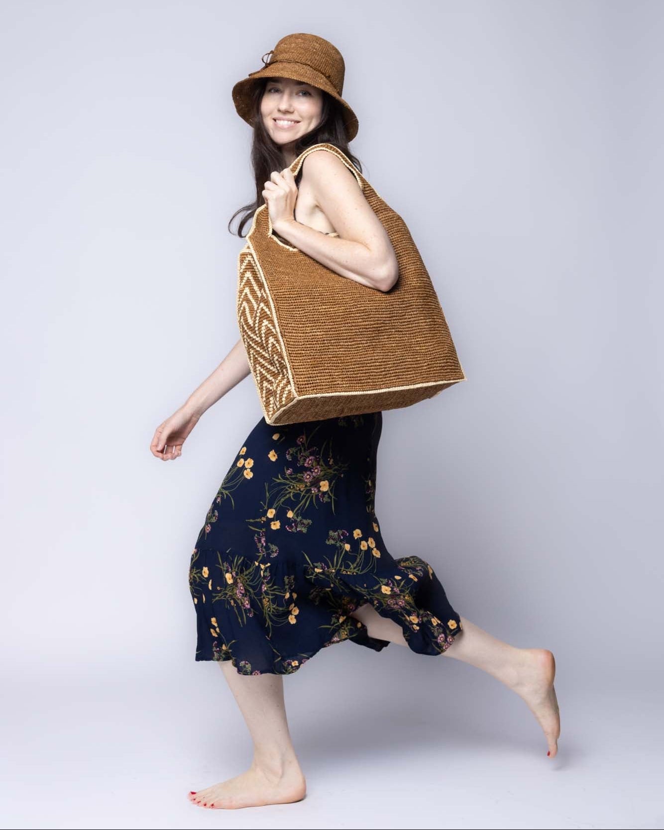 Model wearing Side view Mamie Zigzag Pinstripe XL Crochet Straw Basket Handbag handmade crochet natural raffia palm fiber in a solid hue of tobacco dark/cinnamon/brown purse beach bag with contrasting natural straw color zigzag pinstripe pattern on the sides of the bag and matching outer edge - Shebobo