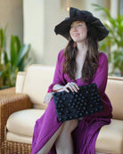 Model wearing Marbella Crochet Straw Oversized Pouch Clutch Purse with Detachable Wristlet Strap handmade crochet natural raffia palm fiber in a solid hue of Black with subtle diamond boucle nub pattern protruding from horizontal bands of link texture small bag - Shebobo (Lilly 5" Brim Fan Pleated Straw Sun Hats with Big Bow)