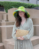 Model wearing Marbella Crochet Straw Oversized Pouch Clutch Purse with Detachable Wristlet Strap handmade crochet natural raffia palm fiber in a solid hue of Natural straw color with subtle diamond boucle nub pattern protruding from horizontal bands of link texture small bag - Shebobo (with Monica 5" & 7" Wide Brim Multicolor Stripe Pattern Packable Straw Sun Hats)
