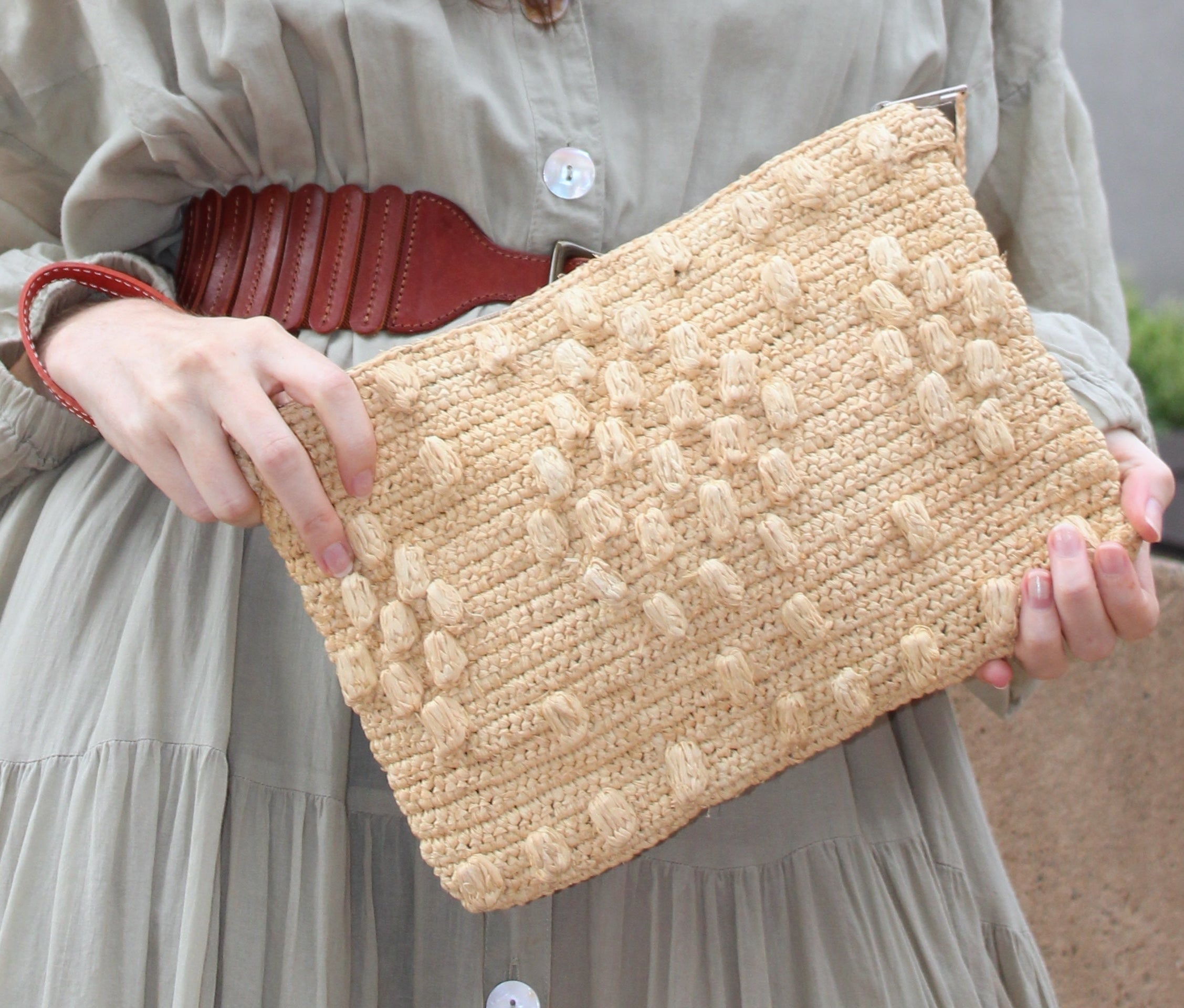 Model wearing Marbella Crochet Straw Oversized Pouch Clutch Purse with Detachable Wristlet Strap handmade crochet natural raffia palm fiber in a solid hue of Natural straw color with subtle diamond boucle nub pattern protruding from horizontal bands of link texture small bag - Shebobo