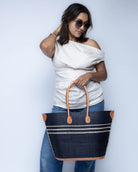 Model wearing Marina Racing Stripe Small Straw Tote Bag handmade loomed natural raffia palm fiber in a solid hue of black with three horizontal bands of natural straw color making a pinstripe pattern across the top third of the bag with matching cross-stitch edging, leather handles and accents beach bag handbag - Shebobo 