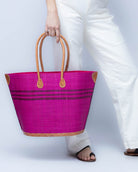 Model wearing Marina Racing Stripe Small Straw Tote Bag handmade loomed natural raffia palm fiber in a solid hue of Fuchsia hot/bright/barbie/pink with three horizontal bands of umberwine burgundy/dark/red/purple making a pinstripe pattern across the top third of the bag with matching cross-stitch edging, leather handles and accents beach bag handbag - Shebobo 
