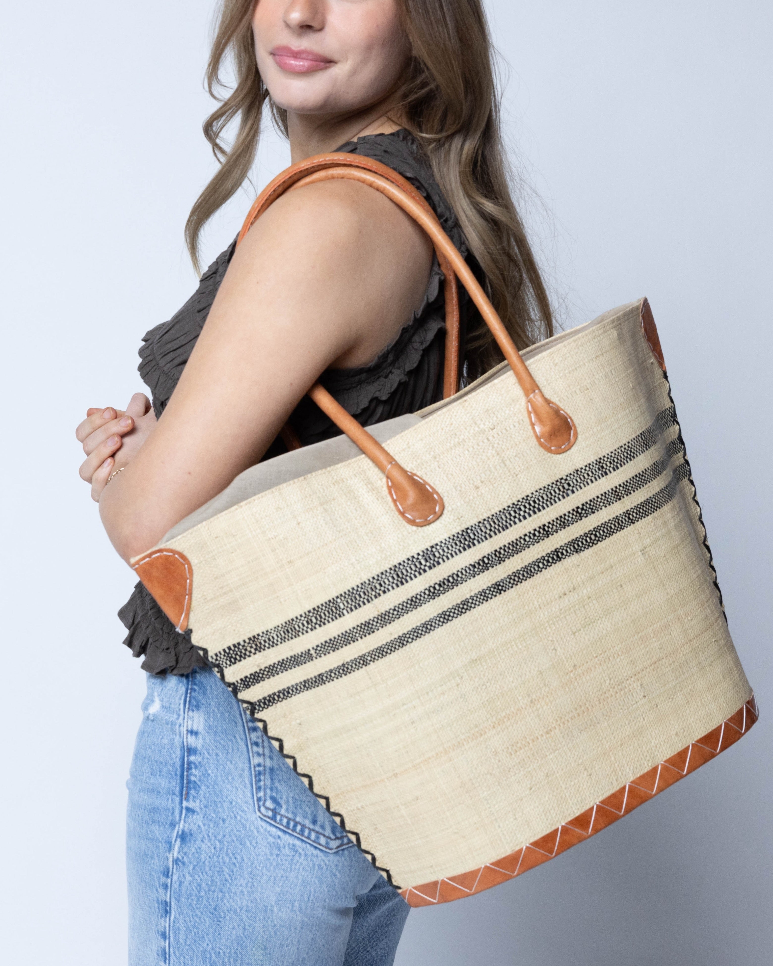 Model wearing Marina Racing Stripe Small Straw Tote Bag handmade loomed natural raffia palm fiber in a solid hue of natural straw color with three horizontal bands of black making a pinstripe pattern across the top third of the bag with matching cross-stitch edging, leather handles and accents beach bag handbag - Shebobo 