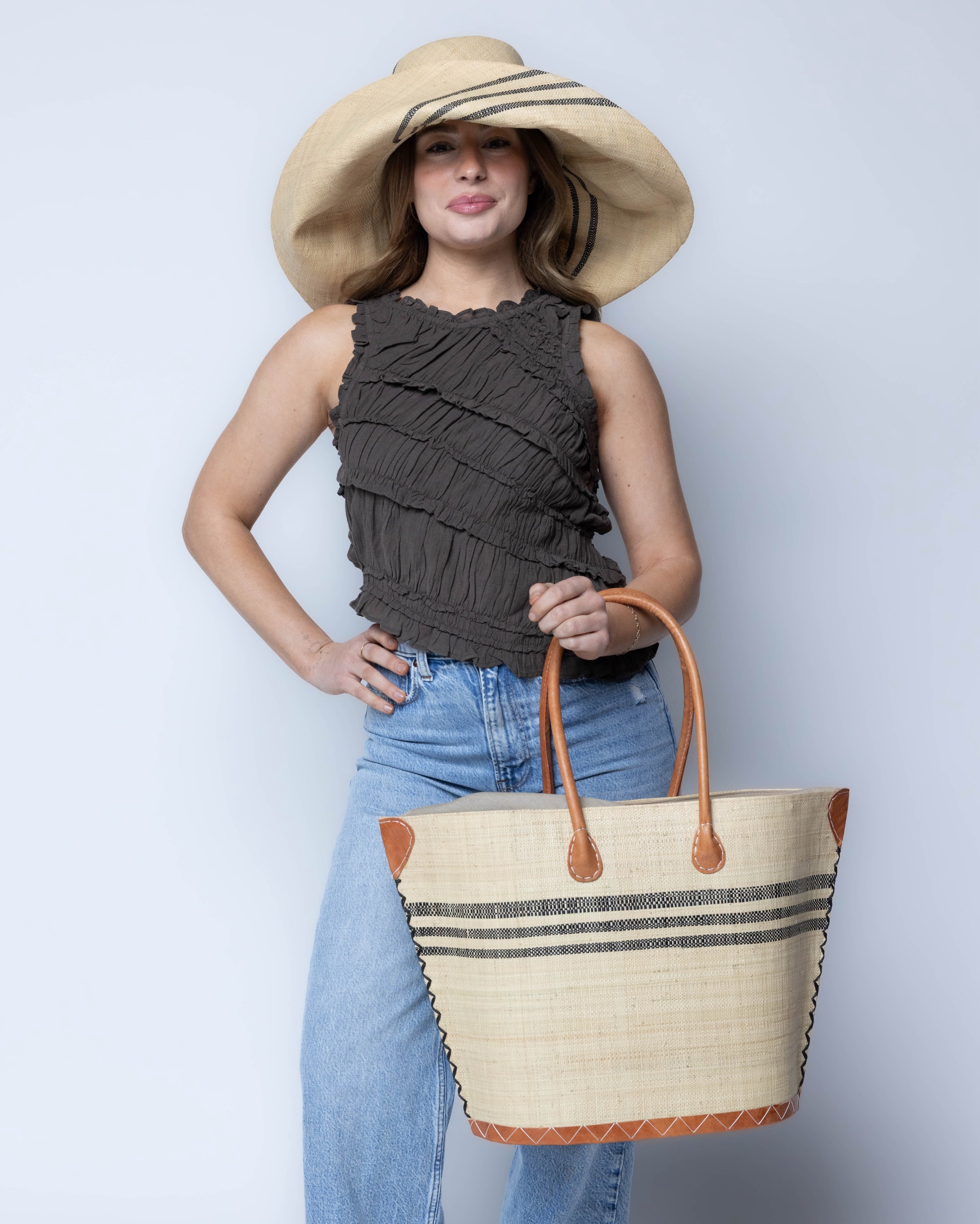 Model wearing Marina Racing Stripe Small Straw Tote Bag handmade loomed natural raffia palm fiber in a solid hue of natural straw color with three horizontal bands of black making a pinstripe pattern across the top third of the bag with matching cross-stitch edging, leather handles and accents beach bag handbag - Shebobo (with Monica 5" & 7" Wide Brim Racing Stripe Packable Straw Sun Hats)