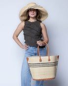 Model wearing Marina Racing Stripe Small Straw Tote Bag handmade loomed natural raffia palm fiber in a solid hue of natural straw color with three horizontal bands of black making a pinstripe pattern across the top third of the bag with matching cross-stitch edging, leather handles and accents beach bag handbag - Shebobo (with Monica 5" & 7" Wide Brim Racing Stripe Packable Straw Sun Hats)