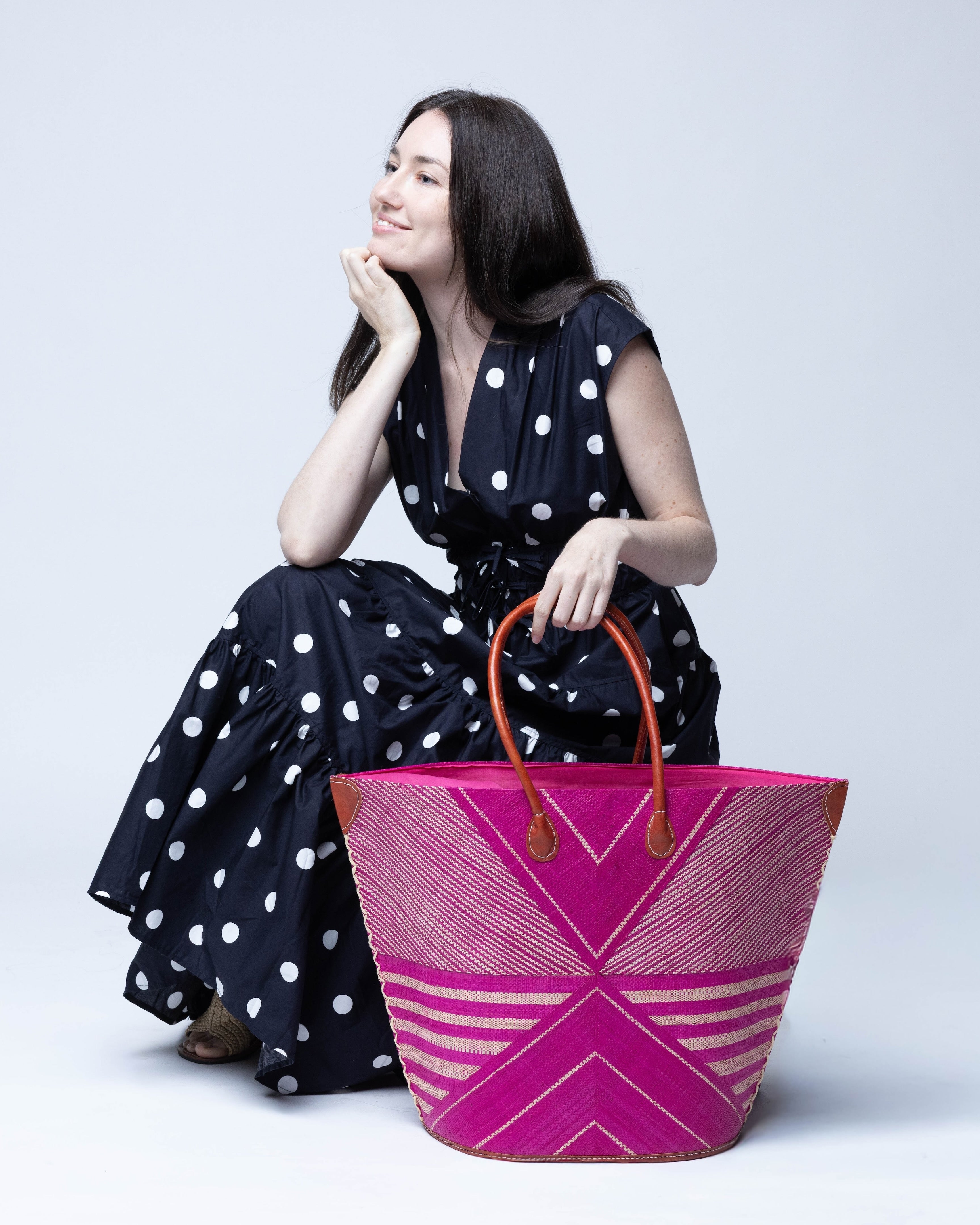 Model wearing Mariposa Large Straw Tote Bag handmade loomed natural raffia palm fiber in a multi paneled pattern design of pinstripe, melange, and wide stripe combinations geometrically radiating from the center point of the bag outwards in a two tone color combination of Fuchsia hot/bright/barbie/pink and natural straw color with a solid hue of color on the back of the bag plus leather handles and embellishments shoulder bag - Shebobo