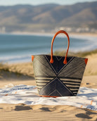 Lifestyle view Mariposa Large Straw Tote Bag handmade loomed natural raffia palm fiber in a multi paneled pattern design of pinstripe, melange, and wide stripe combinations geometrically radiating from the center point of the bag outwards in a two tone color combination of Black and natural straw color with a solid hue of color on the back of the bag plus leather handles and embellishments shoulder bag - Shebobo