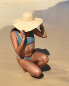 Model wearing Melanie 4.5" Wide Brim Raffia Straw Sun Hats handmade loomed natural raffia palm fiber in a solid hue of natural straw color round crown and flat brim with matching hatband and double bow detailing light weight breathable beach hat - Shebobo 