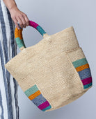 Model wearing Milos Caribbean Stripes Crochet Straw Basket Handbag with Pockets handmade natural raffia palm fiber in a solid hue of natural straw color on the body of the bag with multicolor wide stripe pattern of turquoise blue/green, fuchsia hot/bright/barbie/pink, denim retro/light/blue, saffron yellow/orange, and seafoam light/blue/green on both handles as well as two large external pockets tote bag - Shebobo
