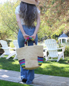 Model wearing Milos Riviera Stripes Crochet Straw Basket Handbag with Pockets handmade natural raffia palm fiber in a solid hue of natural straw color on the body of the bag with multicolor wide stripe pattern of umberwine bordeaux/claret/deep/dark/red/purple, denim retro/light/blue, fuchsia hot/bright/barbie/pink, teal dark/blue/green, midori lime/neon/green, and black on both handles as well as two large external pockets tote bag - Shebobo