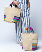 Model wearing Milos Caribbean and Riviera Stripes Crochet Straw Basket Handbag with Pockets handmade natural raffia palm fiber in a solid hue of natural straw color on the body of the bag with multicolor wide stripe pattern on both handles as well as two large external pockets tote bag - Shebobo
