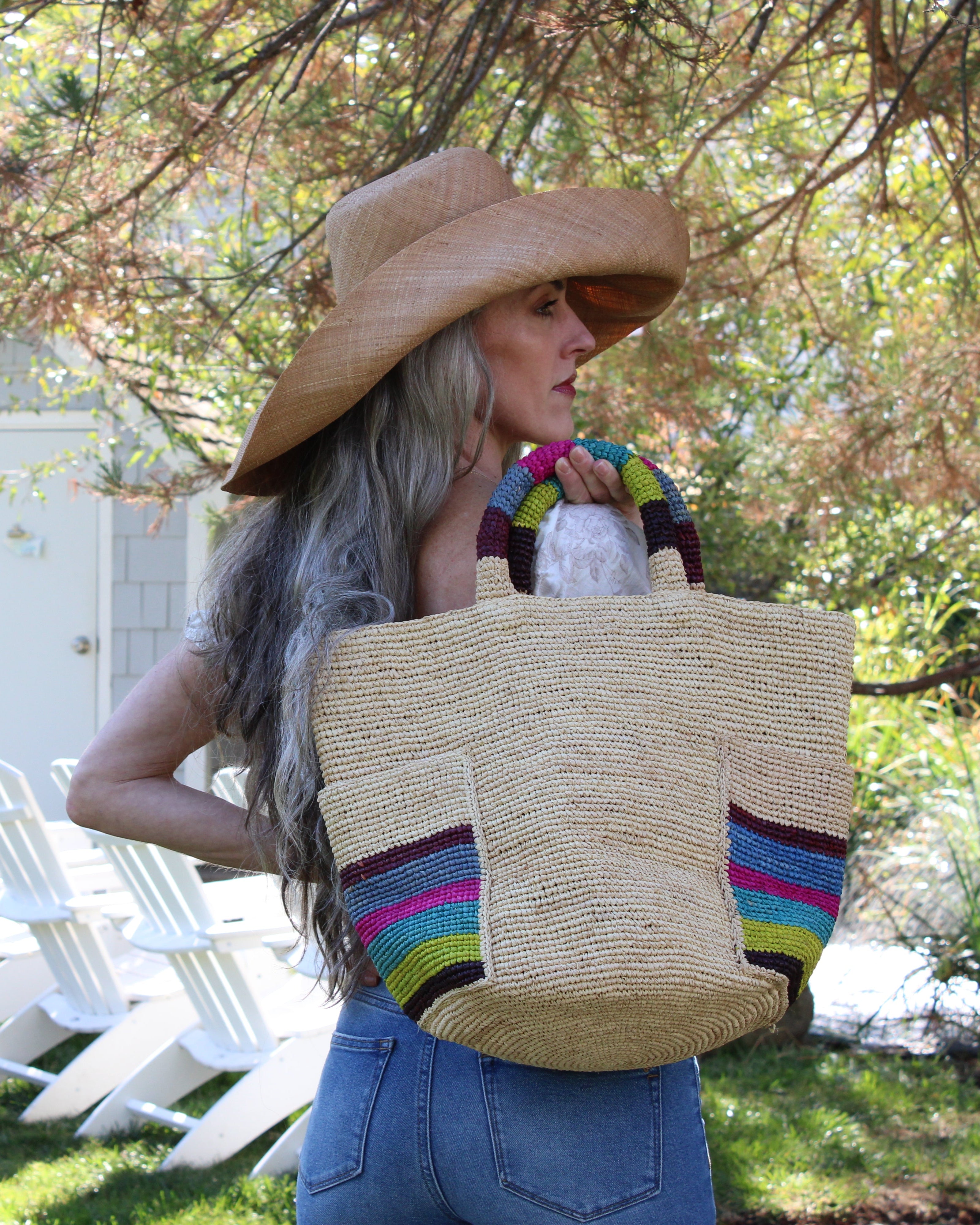 Model wearing Milos Riviera Stripes Crochet Straw Basket Handbag with Pockets handmade natural raffia palm fiber in a solid hue of natural straw color on the body of the bag with multicolor wide stripe pattern of umberwine bordeaux/claret/deep/dark/red/purple, denim retro/light/blue, fuchsia hot/bright/barbie/pink, teal dark/blue/green, midori lime/neon/green, and black on both handles as well as two large external pockets tote bag - Shebobo (with Audrey Wide Brim Big Bow Solid Color Packable Straw Sun Hat)