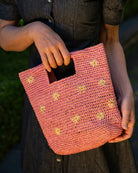 Model wearing Mini ConCon Flowers Crochet Petite Straw Basket handmade woven raffia palm fiber in a solid hue of pink making a textured link pattern with delicate hand embroidered daisy floral pattern of natural straw color with dot of saffron orange/yellow in the center of each flower randomly placed around the entire bag handbag purse small bag - Shebobo