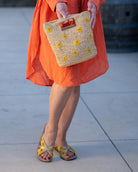 Model wearing Daisy Crochet Straw & Leather Sandal with Flower Embroidery & Metallic Detailing handmade crochet natural raffia palm fiber in a solid hue of natural straw color in two bands with saffron yellow petals and coral orange/red centered embroidered flower pattern wrap around the top  of the shoe with a band of metallic gold across the top summer sandals shoes - Shebobo (with Mini Concon Flower Embroidery Straw Basket Purse)