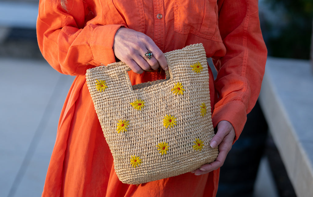 Model wearing Mini ConCon Flowers Crochet Petite Straw Basket handmade woven raffia palm fiber in a solid hue of natural straw color making a textured link pattern with delicate hand embroidered daisy floral pattern of saffron yellow with dot of coral orange/red in the center of each flower randomly placed around the entire bag handbag purse small bag - Shebobo