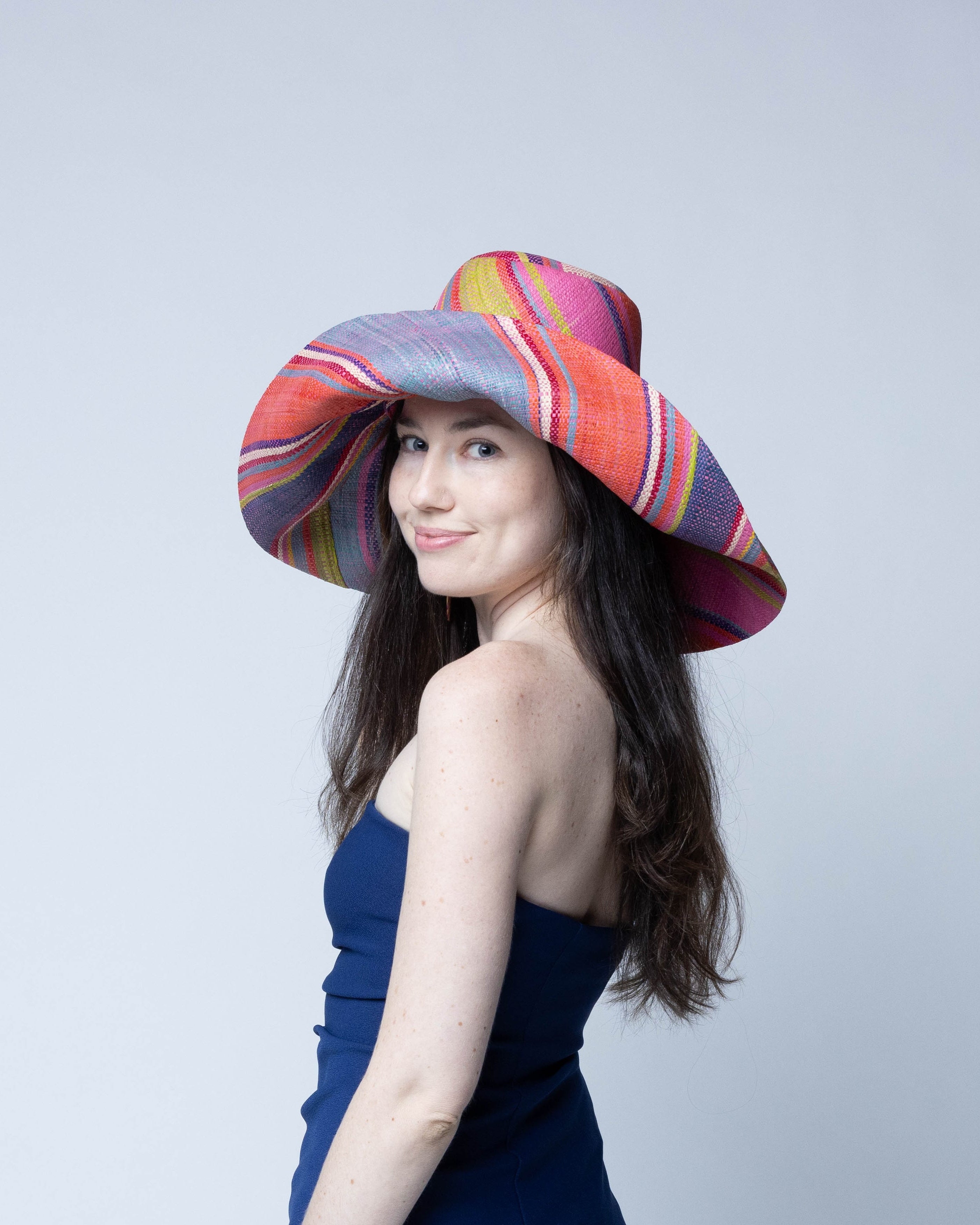 Model wearing Monica 7" Wide Brim Raspberry Swirl Multicolor Stripes Packable Straw Sun Hat Handmade loomed raffia in multi width bands of purple, light pink, coral orange/red, lime green, natural straw color, light blue, yellow, etc. create a swirl pattern - Shebobo