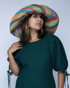 Model wearing Monica 7" Wide Brim Amalfi Swirl Multicolor Stripes Packable Straw Sun Hat handmade loomed raffia in bands of natural straw color, mocha chocolate/dark/brown, green, red, and turquoise blue stripes create a swirl pattern - Shebobo