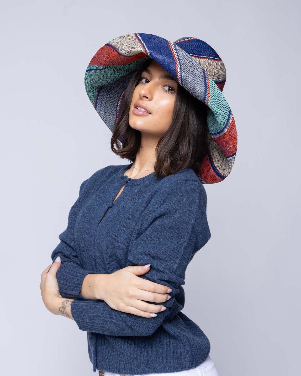 Model wearing Monica 5" & 7" Wide Brim Coral & Blue Swirl Multicolor Stripes Packable Straw Sun Hat Handmade loomed raffia in multi width bands of denim light/blue, navy dark/blue, coral orange/red, seafoam light/blue/green, and natural straw color create a swirl pattern - Shebobo