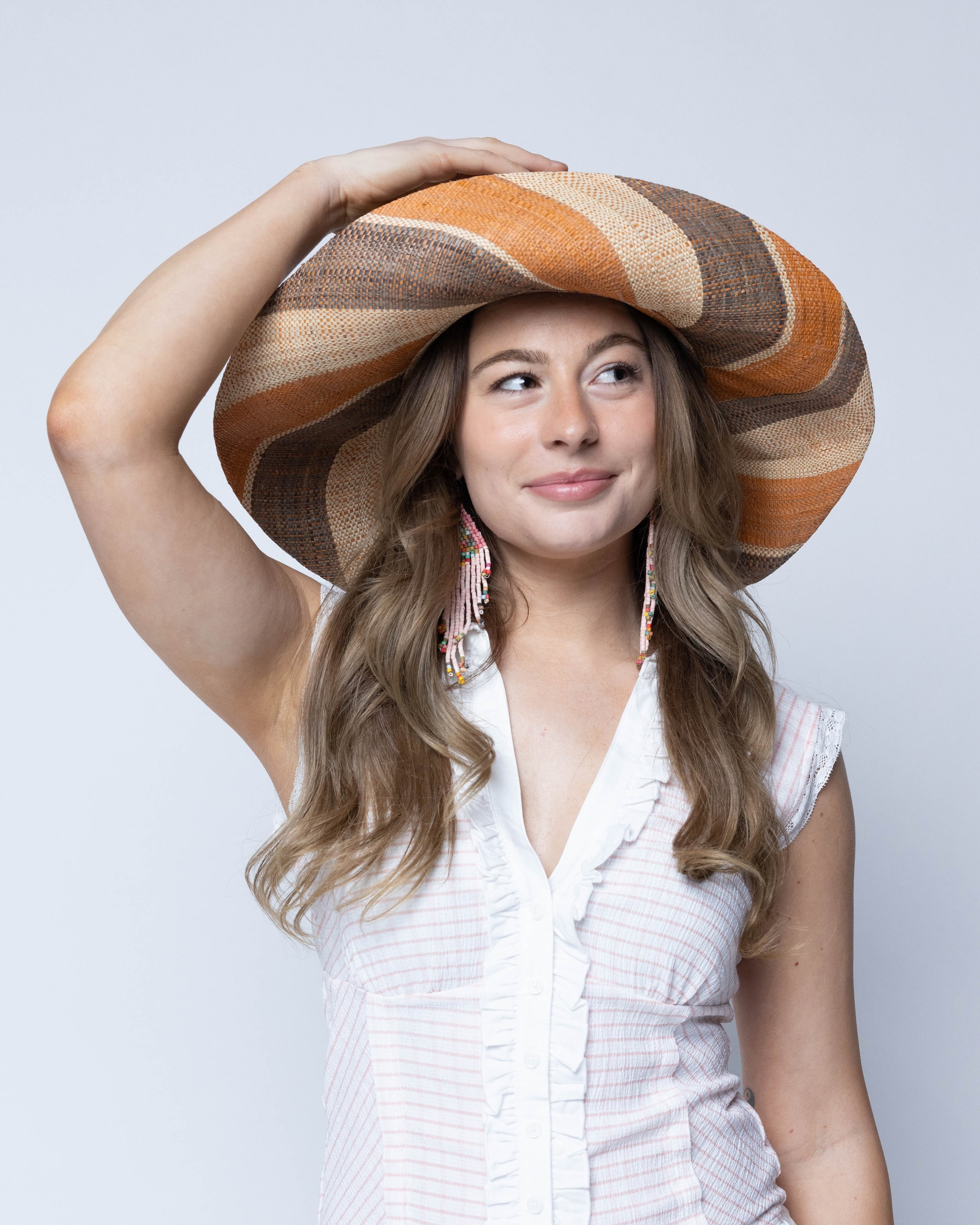 Model wearing Monica 5" Wide Brim Cinnamon Swirl Multicolor Stripe Pattern Packable Straw Sun Hat handmade loomed raffia in multi width bands of cinnamon/tobacco/brown, grey, and natural straw color create a striped swirl pattern - Shebobo