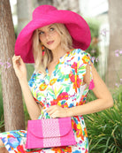 Model wearing Wynwood Fuchsia Straw Handbag with Metallic Detailing & Horn Handle handmade loomed raffia in fuchsia pink/hot pink/barbie pink and silver metallic vegan leather in three vertical bands of color with the metallic weave centered on the purse under the handles with Monica 7" Wide Brim Packable Straw Sun Hat - Shebobo