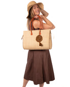 Model wearing Monte Carlo Big Straw Beach Bag with Dreamcatcher Tassel Charm Embellishment handmade loomed raffia palm fiber in a solid hue of natural straw color with contrasting crosshatch stitch edging in cinnamon/tobacco/dark/brown with matching crochet dreamcatcher disc and tuft tassel charm embellishment and leather handles oversized large handbag shoulder bag - Shebobo (with 4" Brim Genevieve Crochet Straw Sun Hat with Lace Weave Edge)