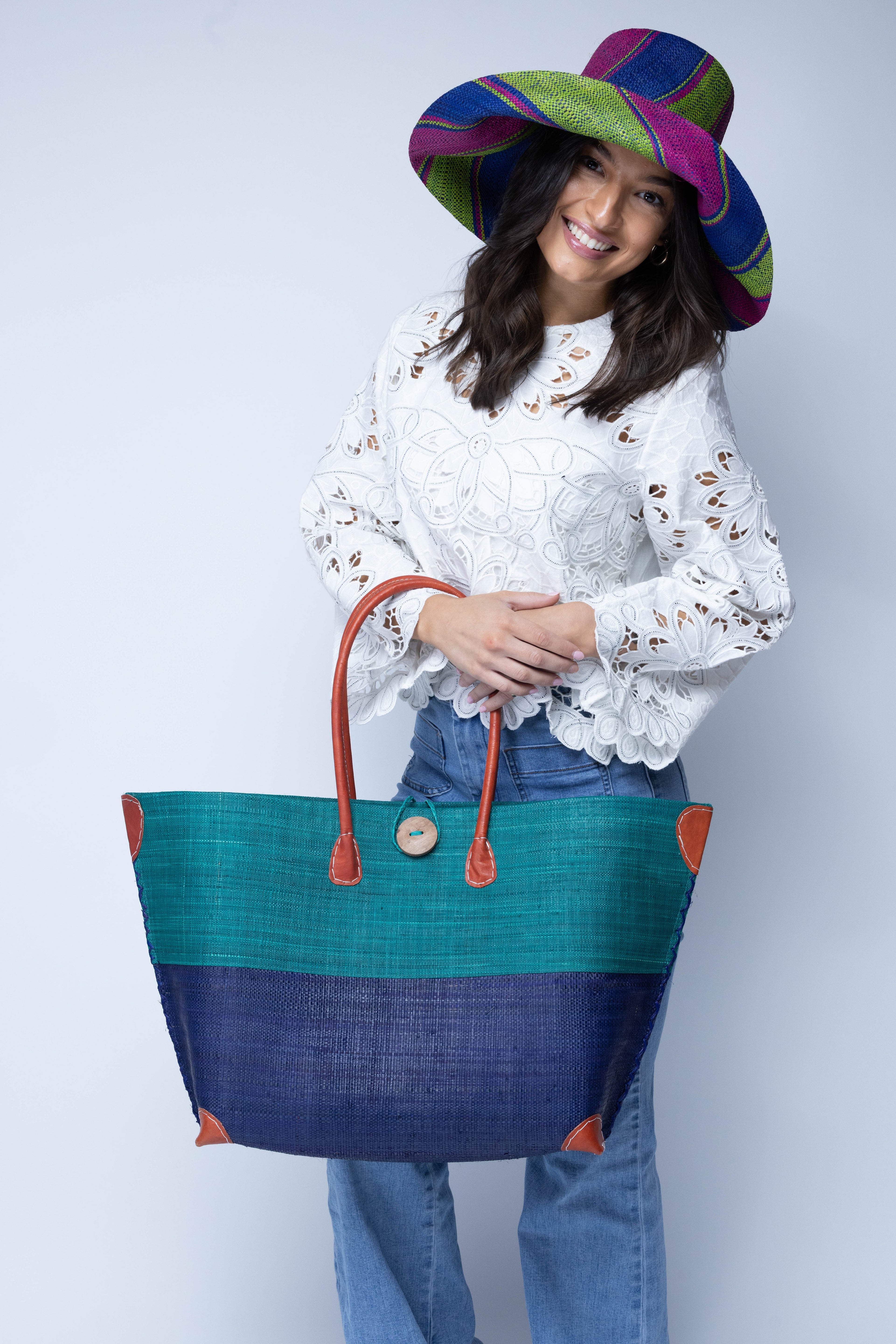 Model wearing Monterey Two Tone Teal and Dusk Straw Tote Bag handmade loomed raffia fibers in a color block pattern of teal dark/blue/green colored upper half, and dusk dark/navy/blue lower half - Shebobo (with Monica 5" & 7" Wide Brim Multicolor Stripe Pattern Packable Straw Sun Hats)