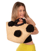 Model wearing Phi Phi Black and Natural Braided Straw Basket with Oversized Raffia Poufs handmade raffia palm fiber braided into a solid hue of natural straw color with three randomly placed black hand-brushed fringe poufs per side for embellishment with matching loop wrap top edging and matching crochet loop/braided handles bag handbag - Shebobo