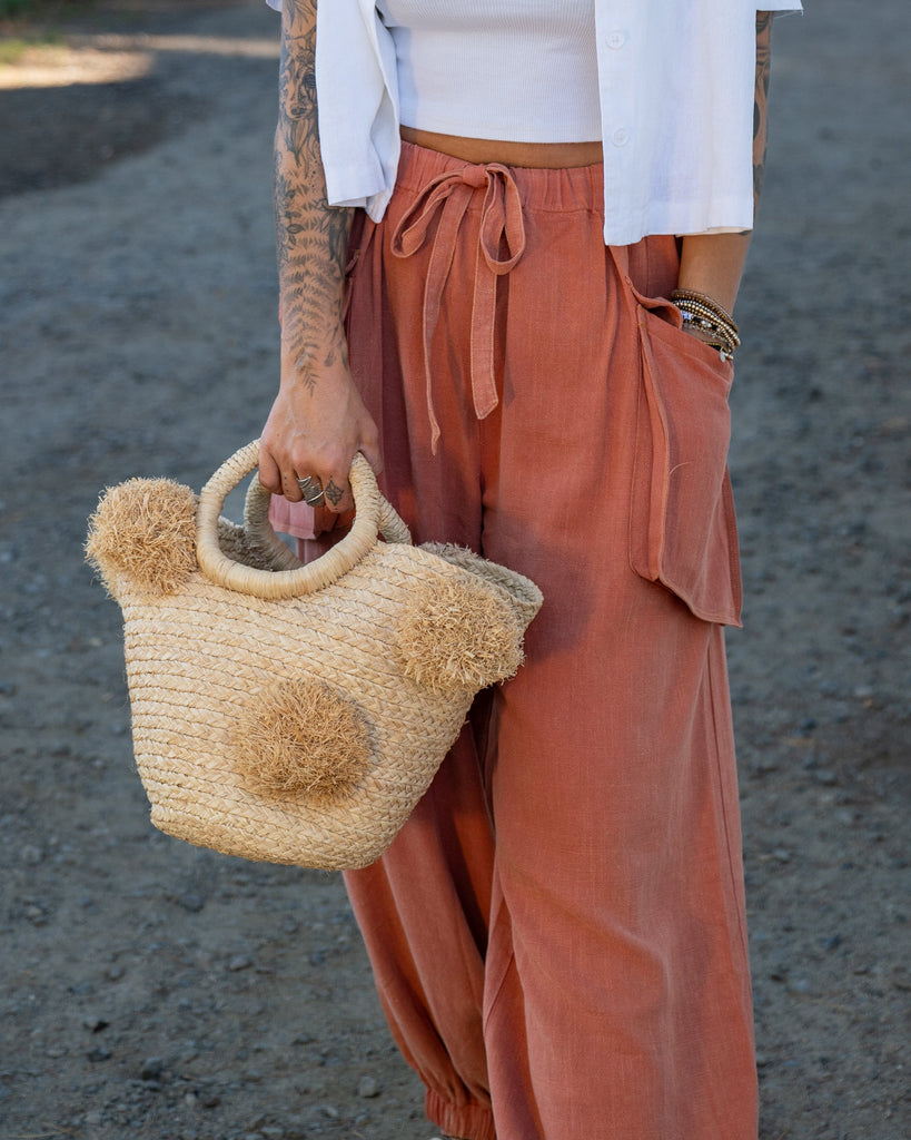 Model wearing Phi Phi  Natural Braided Straw Basket with Oversized Raffia Poufs handmade raffia palm fiber braided into a solid hue of natural straw color with three randomly placed matching hand-brushed fringe poufs per side for embellishment with loop wrap top edging and crochet loop/braided handles bag handbag - Shebobo