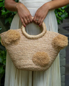 Model wearing Phi Phi  Natural Braided Straw Basket with Oversized Raffia Poufs handmade raffia palm fiber braided into a solid hue of natural straw color with three randomly placed matching hand-brushed fringe poufs per side for embellishment with loop wrap top edging and crochet loop/braided handles bag handbag - Shebobo