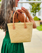 Model wearing Pianina Natural Small Straw Basket Bag handmade from woven natural raffia palm fiber in a monochromatic cross weave pattern of natural straw color handbag purse with leather handles - Shebobo