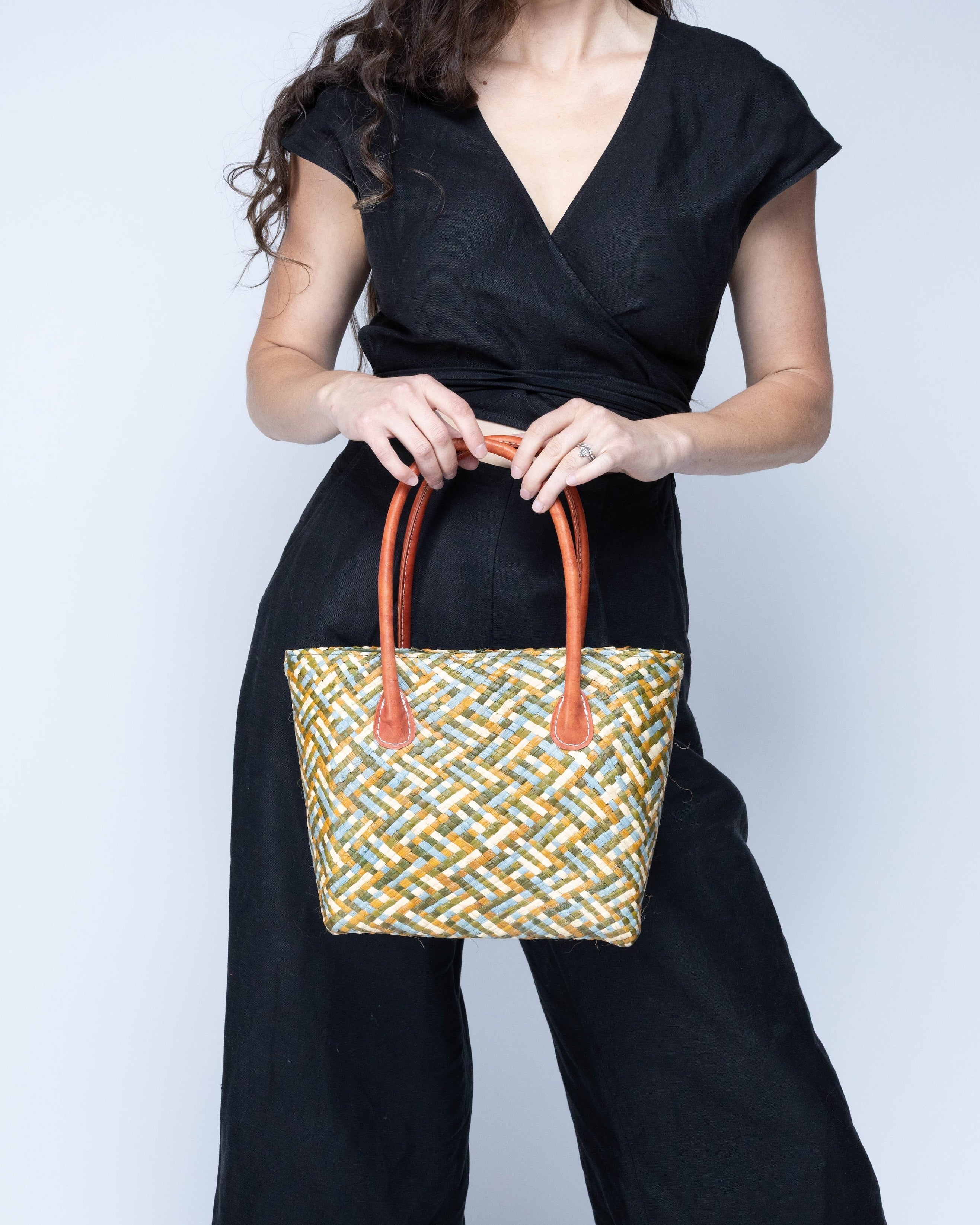 Model wearing Pianina Terra Multi Small Straw Basket Bag handmade from woven natural raffia palm fiber in a multicolor cross weave pattern of dark green, olive green, denim light/blue, and natural straw color handbag purse with leather handles - Shebobo