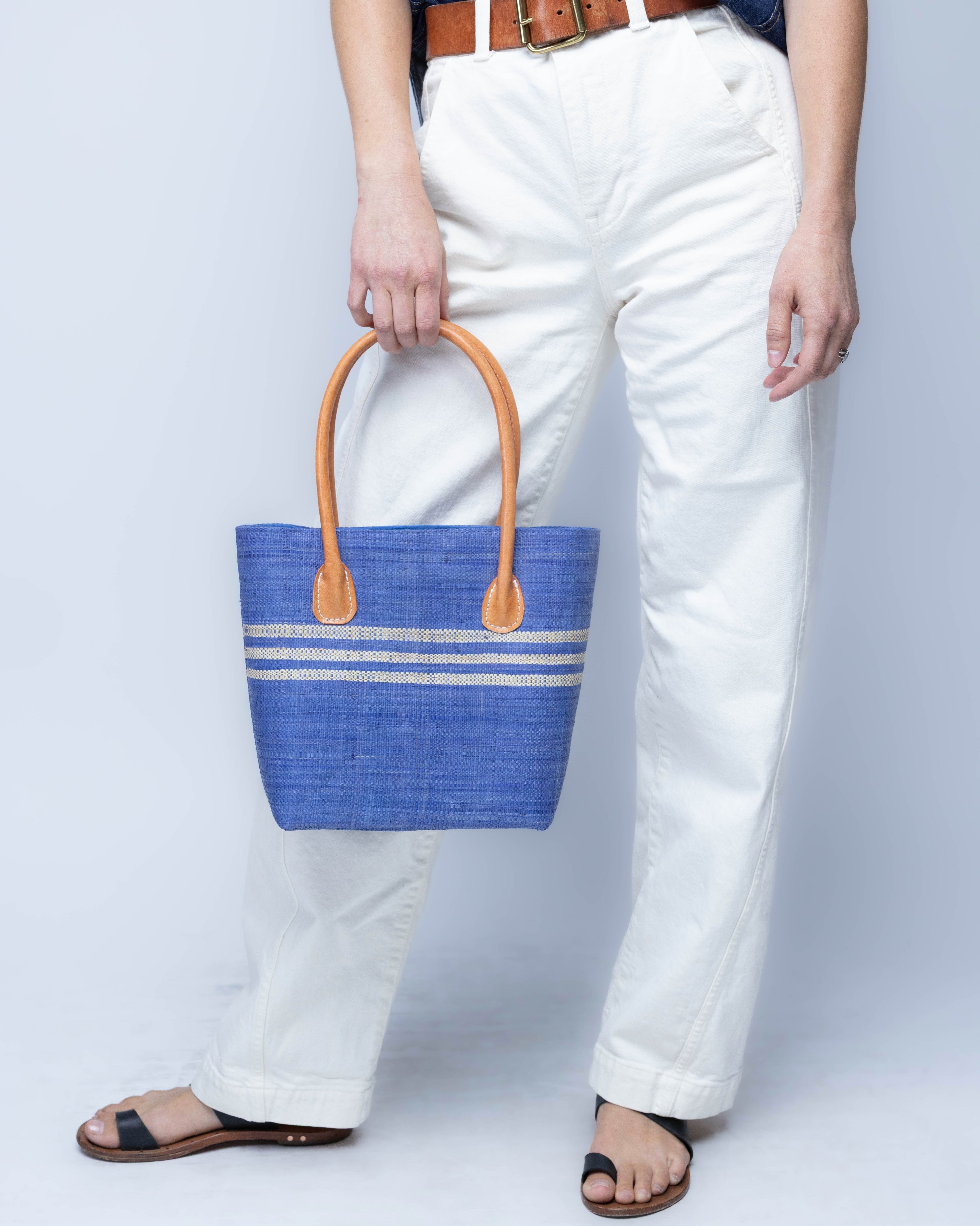 Model wearing Raja Racing Stripe Straw Basket Bag handmade loomed natural raffia palm fiber in a two tone multicolor design of Denim light/retro/soft/blue color with three horizontal bands of Natural straw color creating a racing stripe pattern placed under the base of the handles a third down the height of the bag - Shebobo