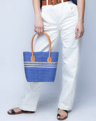 Model wearing Raja Racing Stripe Straw Basket Bag handmade loomed natural raffia palm fiber in a two tone multicolor design of Denim light/retro/soft/blue color with three horizontal bands of Natural straw color creating a racing stripe pattern placed under the base of the handles a third down the height of the bag - Shebobo