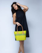 Model wearing Raja Racing Stripe Straw Basket Bag handmade loomed natural raffia palm fiber in a two tone multicolor design of Midori lime/bright/neon/green color with three horizontal bands of Turquoise blue/green creating a racing stripe pattern placed under the base of the handles a third down the height of the bag - Shebobo