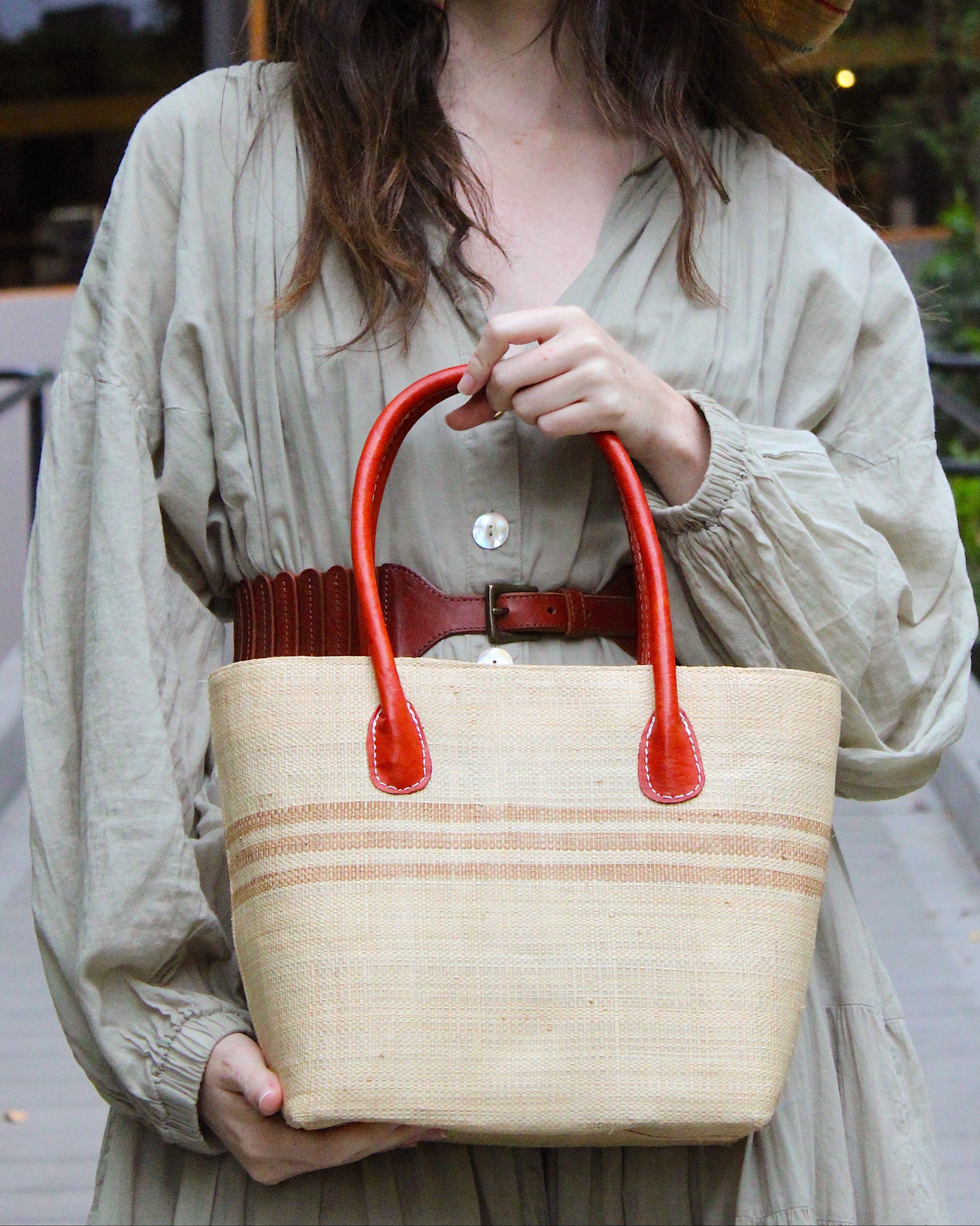 Model wearing Raja Racing Stripe Straw Basket Bag handmade loomed natural raffia palm fiber in a two tone multicolor design of Natural straw color with three horizontal bands of Cappuccino tea/light/brown creating a racing stripe pattern placed under the base of the handles a third down the height of the bag - Shebobo (with Monica 5" & 7" Wide Brim Multicolor Pinstripe Pattern Packable Straw Sun Hats)