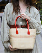 Model wearing Raja Racing Stripe Straw Basket Bag handmade loomed natural raffia palm fiber in a two tone multicolor design of Natural straw color with three horizontal bands of Cappuccino tea/light/brown creating a racing stripe pattern placed under the base of the handles a third down the height of the bag - Shebobo (with Monica 5" & 7" Wide Brim Multicolor Pinstripe Pattern Packable Straw Sun Hats)