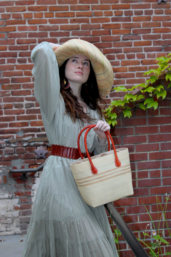 Model wearing Raja Racing Stripe Straw Basket Bag handmade loomed natural raffia palm fiber in a two tone multicolor design of Natural straw color with three horizontal bands of Cappuccino tea/light/brown creating a racing stripe pattern placed under the base of the handles a third down the height of the bag - Shebobo (with Monica 5" & 7" Wide Brim Multicolor Pinstripe Pattern Packable Straw Sun Hats)