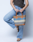 Model wearing Rayo Stripes Straw Basket Bag Tobacco Swirl handmade loomed natural raffia palm fiber in a multicolor multi width horizontal stripe pattern of grey, tobacco cinnamon/dark/brown, denim retro/light/blue and natural straw color with leather handles and accents tote beach bag - Shebobo
