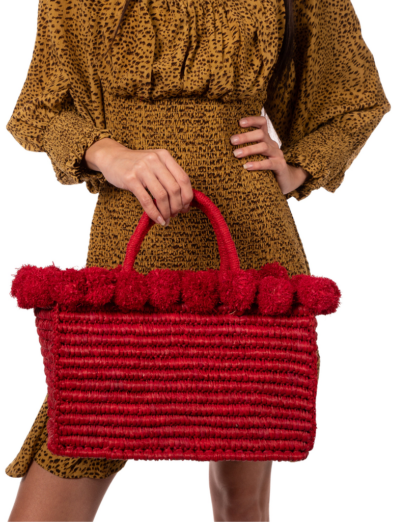 Model wearing Rico Red Crochet Rectangle Straw Basket handmade woven raffia palm fiber in a solid hue of red with horizontal bands of looped texture with matching small raffia pouf pompom top edge and wrapped braid handles small handbag purse - Shebobo