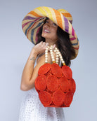Model wearing Romy Coral Crochet Disc Straw Handbag with Wooden Bead Handles handmade natural raffia palm fiber in a solid hue of coral orange/red in a crochet coiled circular flat disc attached together with macrame knot technique with a geometric hexagonal overall shape with seven discs per side two above and below a horizontal row of three per side and a single row comprising the side of the bag with beige carved round wooden beaded handles purse - Shebobo (with Monica 7" Wide Brim Packable Straw Sun Hat